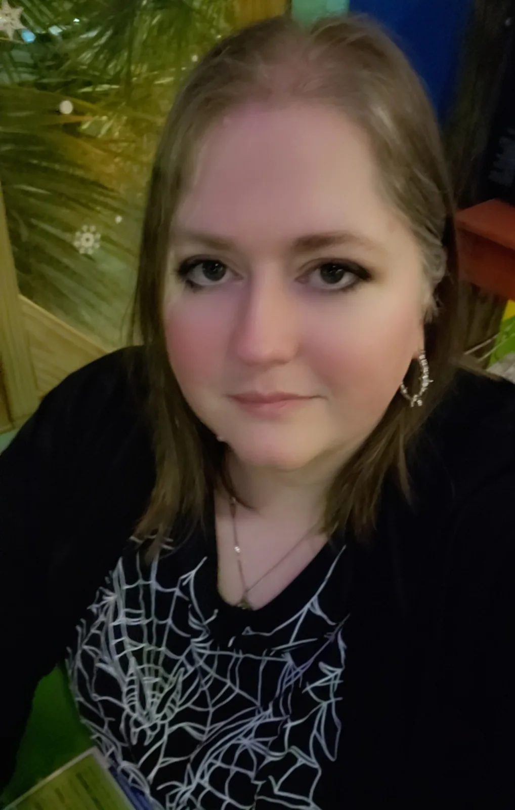 A person with shoulder-length brown hair and dark eye makeup looks directly at the camera with a neutral expression, wearing…