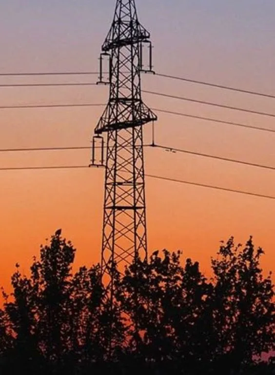 A silhouette of a tall electricity pylon against a vibrant orange and purple sunset sky, with dark tree tops visible at the…