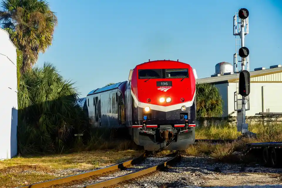 An Amtrak train, numbered 196, is shown approaching on tracks with palm trees and a building in the background. A red signal…