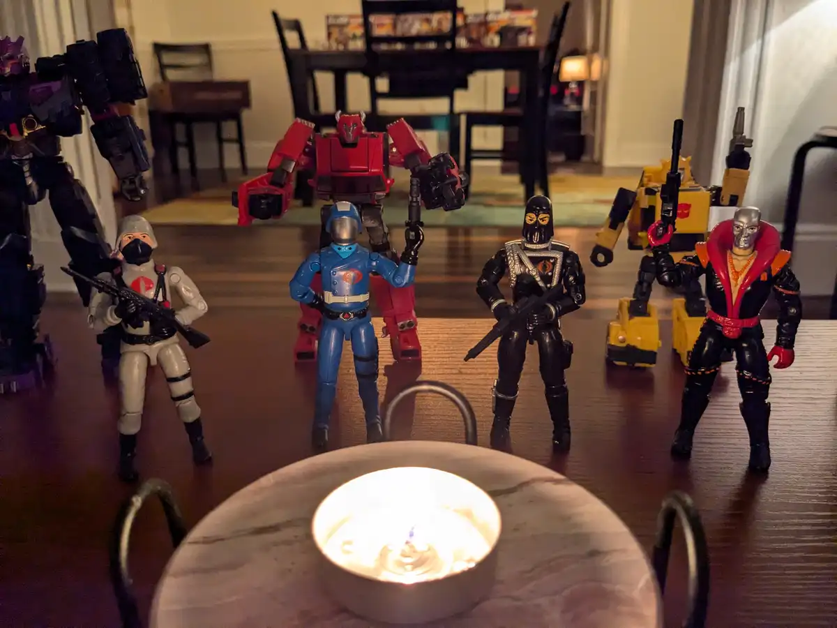 Several G.I. Joe and Transformers action figures are arranged around a lit candle on a table, appearing to be gathered for a…