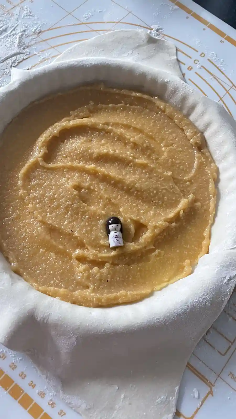 An overhead photo shows a pie crust filled with a thick, yellowish filling, with a tiny figurine of a doll placed in the…