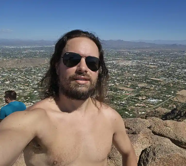 A shirtless man with long hair and sunglasses takes a selfie atop a rocky hill, overlooking a sprawling suburban landscape…