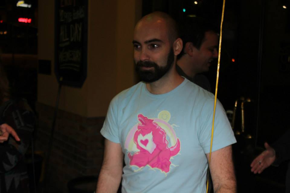 A bald man with a beard wearing a light blue t-shirt featuring a pink, roaring Care Bear with a heart on its chest and a…