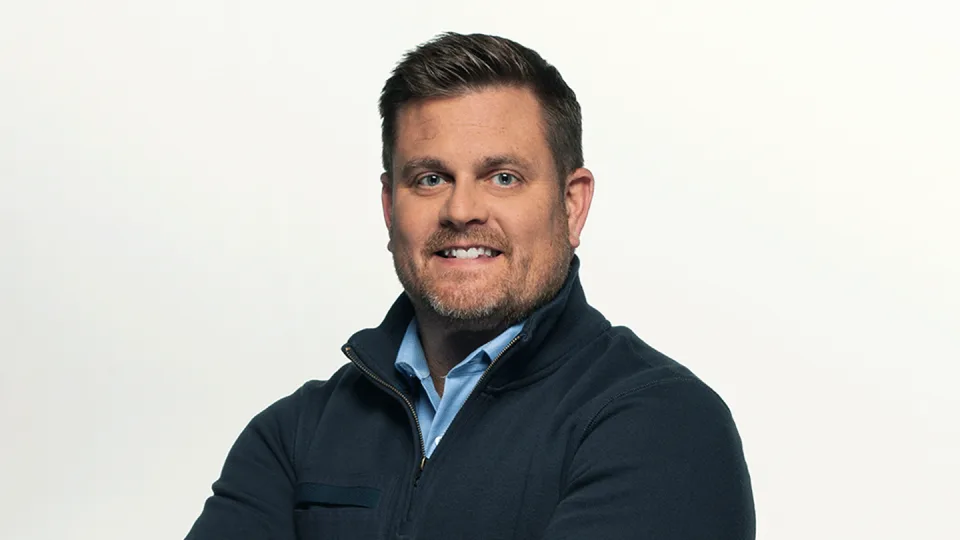 A man with short brown hair and a light beard, wearing a dark blue zip-up jacket over a light blue collared shirt, smiles…