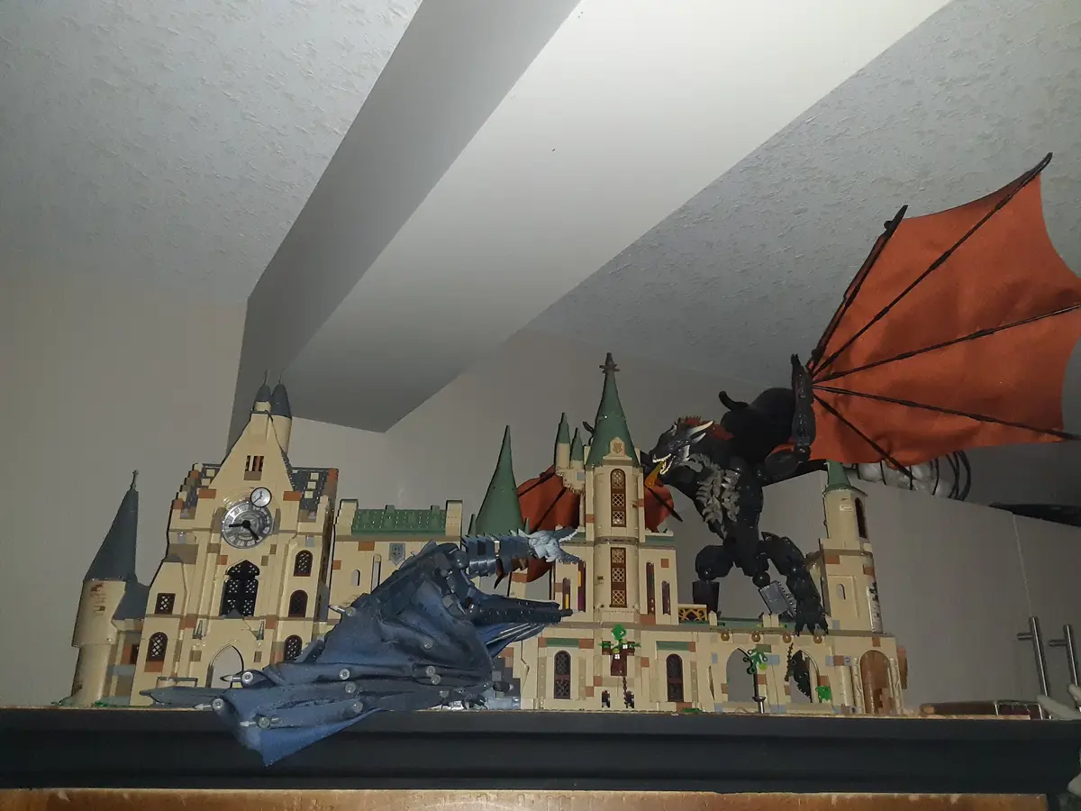 A large LEGO model of Hogwarts Castle is shown with two dragons superimposed on it. One dragon, with orange wings, is…