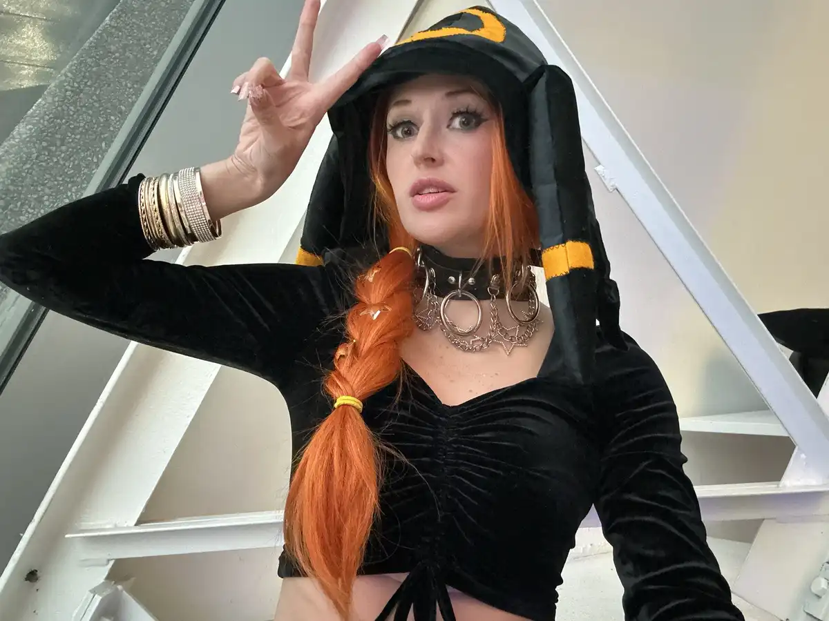 A person with orange hair styled in a braid, wearing a black and yellow penguin-themed hooded costume with ear flaps, poses…