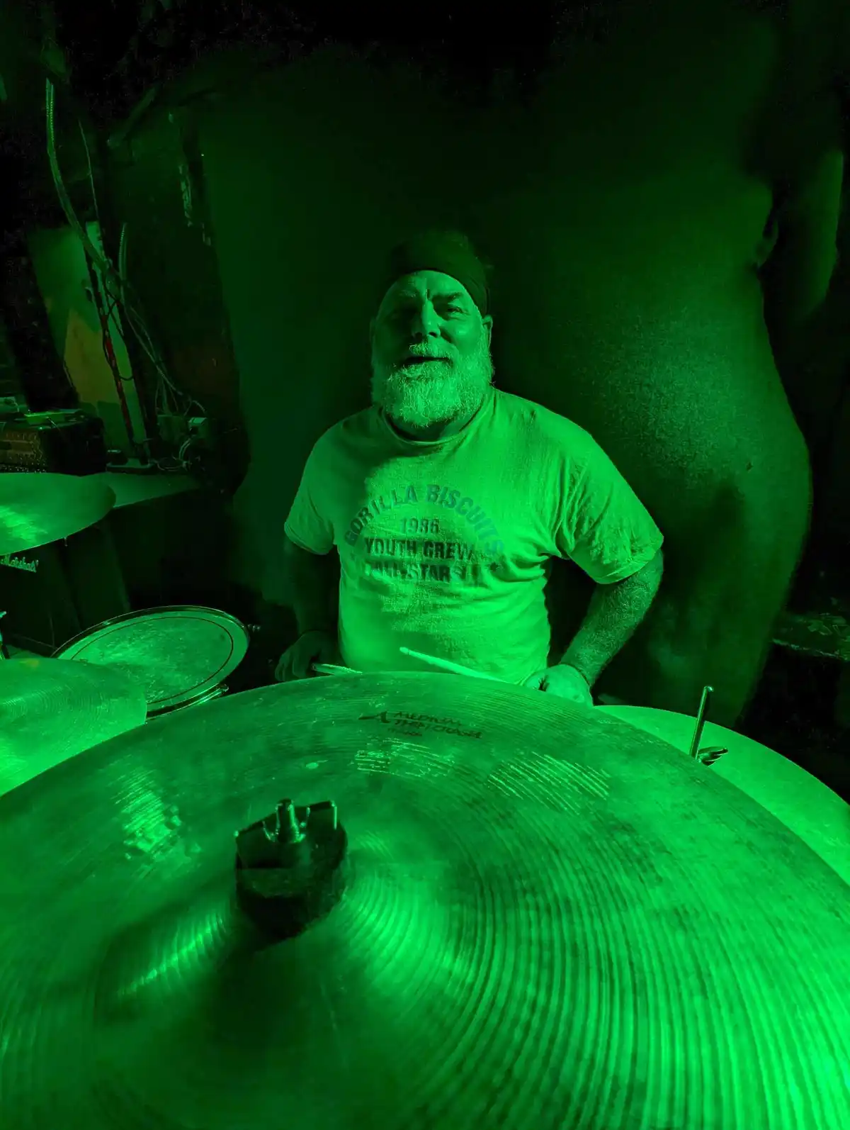 A man with a beard and headband sits behind a drum kit under green stage lighting, wearing a t-shirt that reads 'GORILLA…