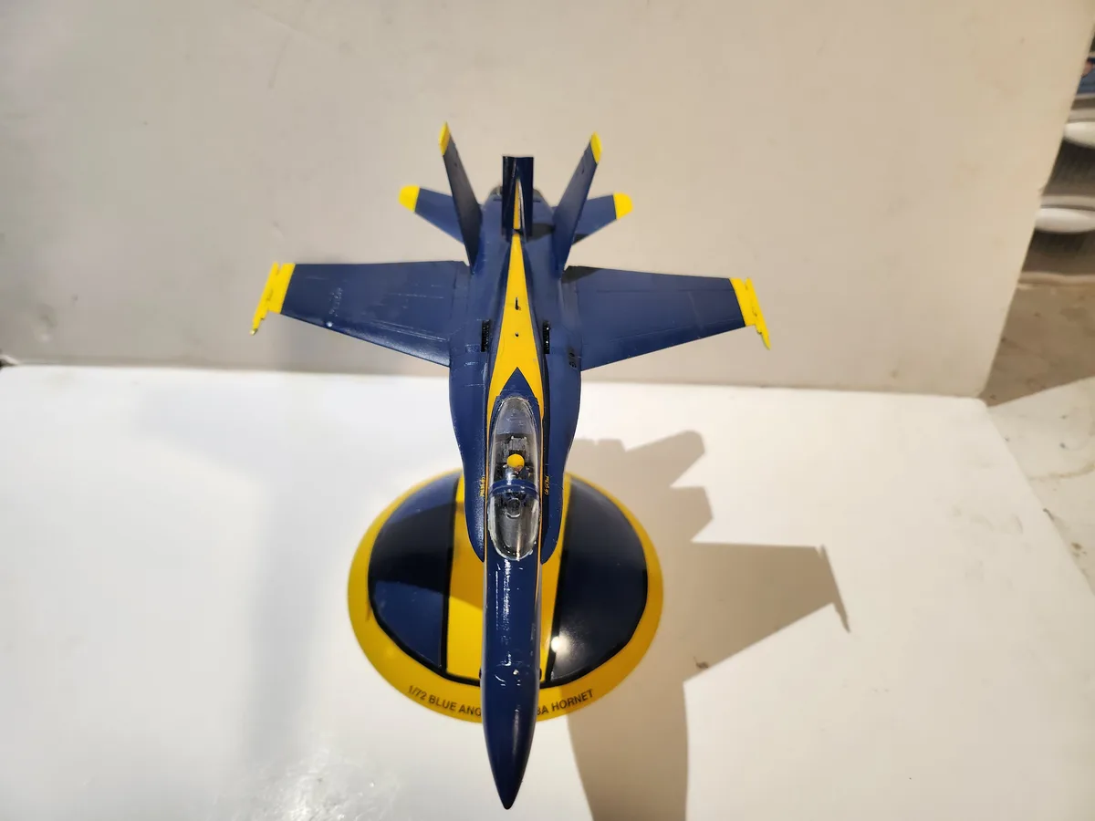 A blue and yellow model of a Blue Angels F/A-18A Hornet jet fighter is displayed from a top-down perspective on a matching…