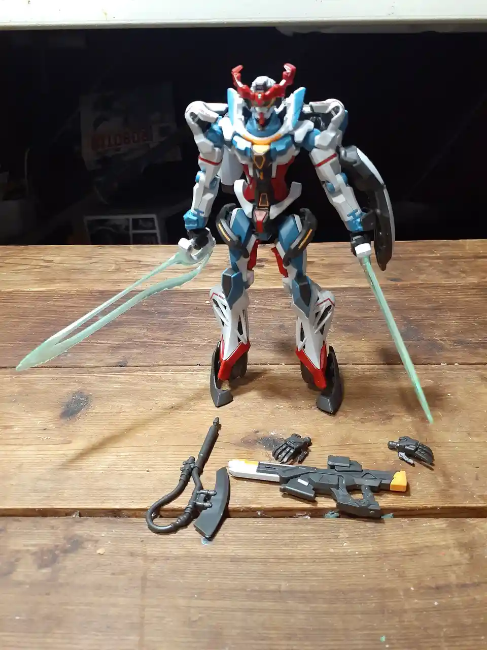 A blue, white, and red mecha action figure stands on a wooden surface holding two translucent green swords, with several…