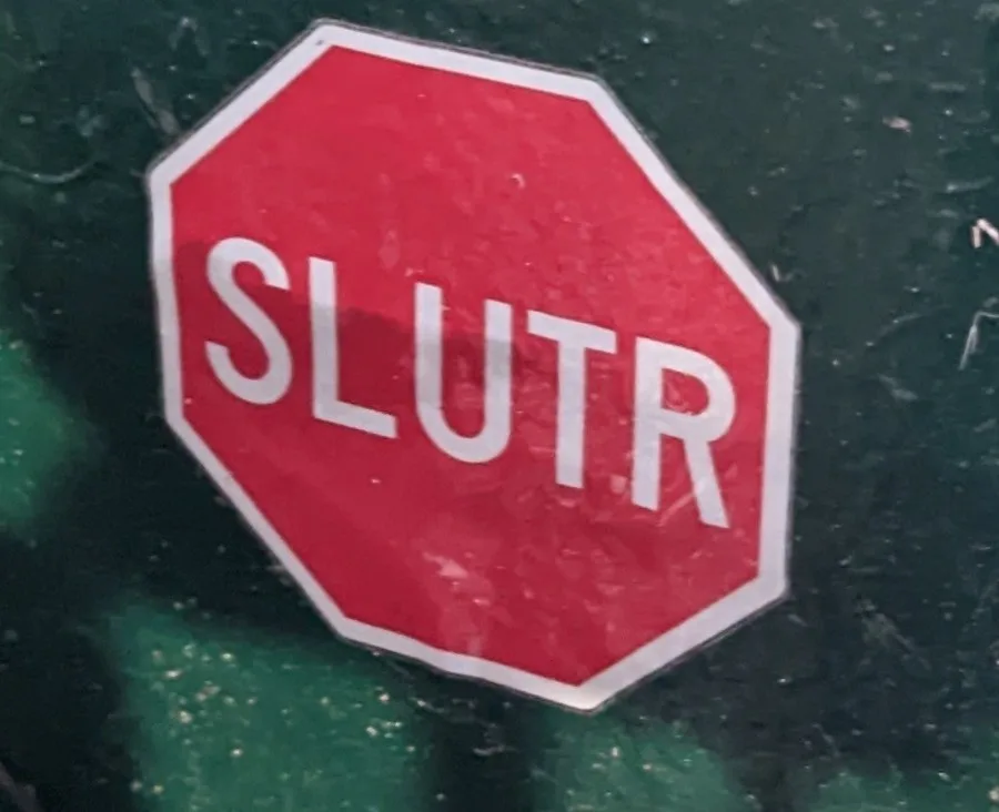A red octagonal stop sign sticker with the word 'SLUTR' written in white capital letters, affixed to a dark green, textured…