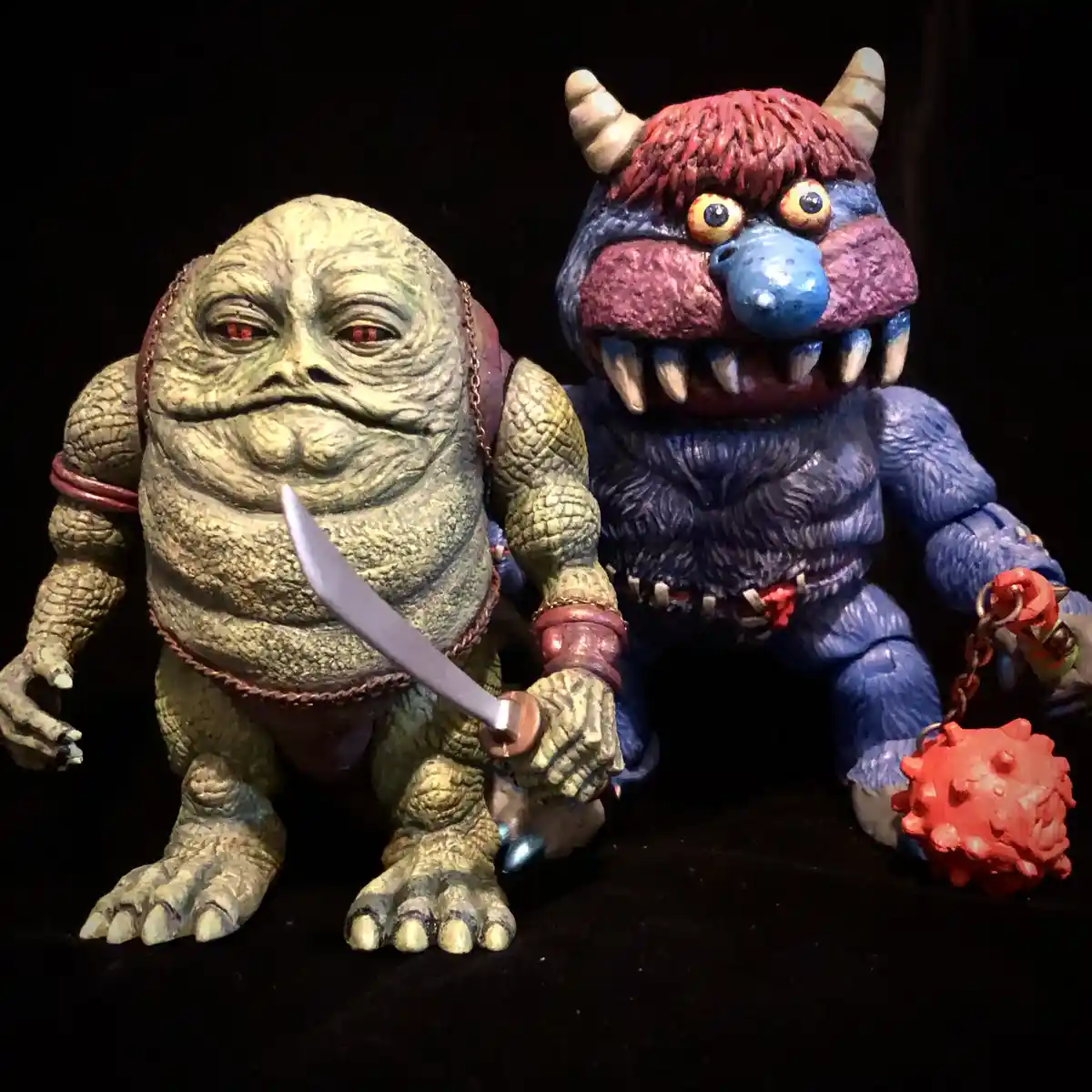 Two action figures are posed side-by-side against a black background. The figure on the left is a green, wrinkled, reptilian…
