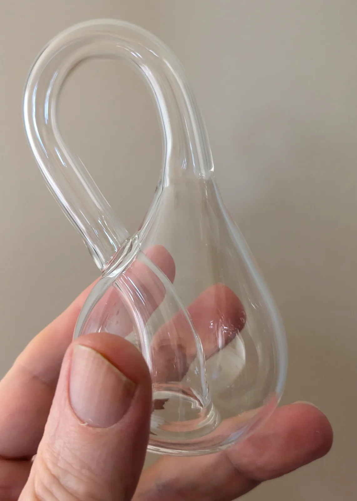 A hand holds a clear, glass object shaped like a stylized, curved dildo with a bulbous base and a looped shaft. The object…
