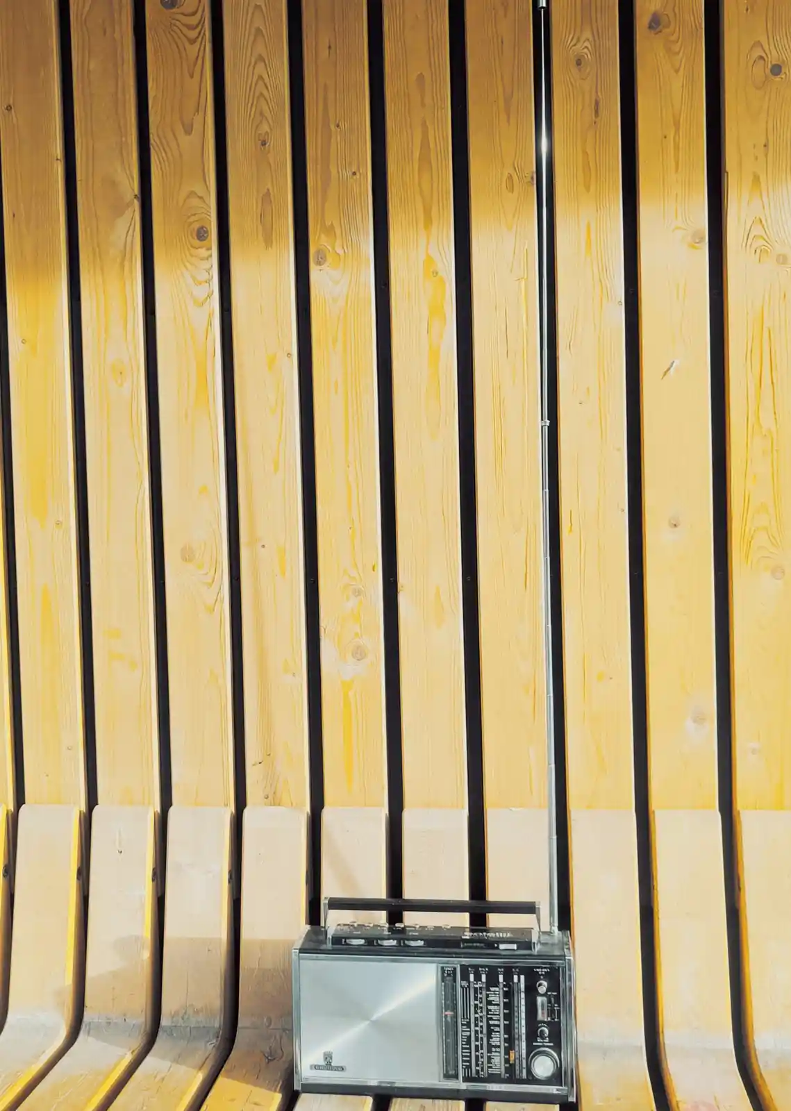 A vintage silver portable radio with an extended antenna sits on a wooden bench with vertical slats, against a backdrop of…