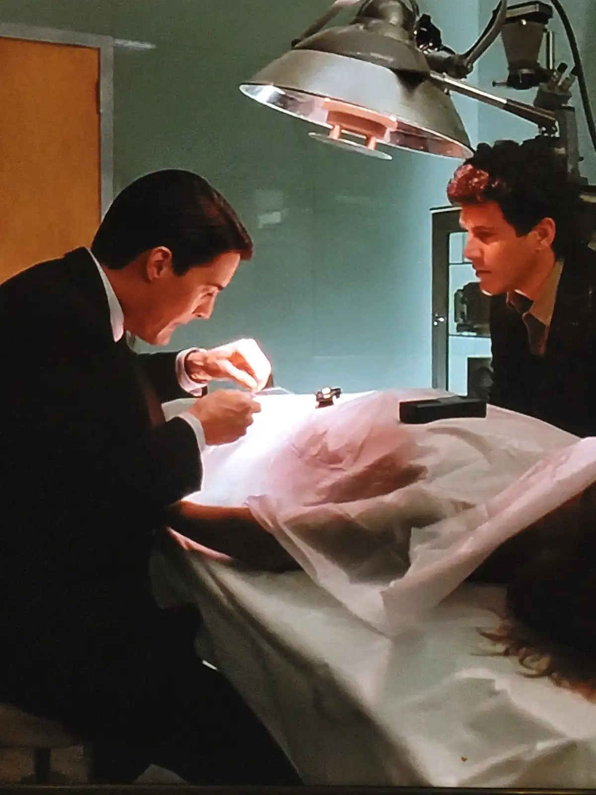 Two men in suits are in a lab or morgue setting, one is intently working on a small object under a bright lamp while the…