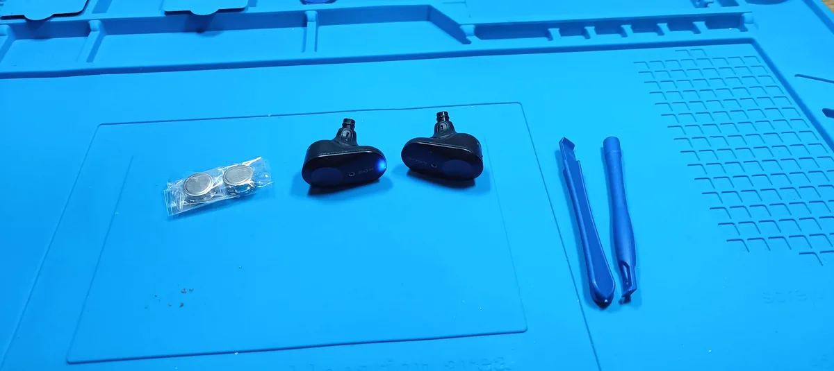 A blue plastic work mat holds two black Sony wireless earbuds, a small plastic bag with two coin cell batteries, and two…