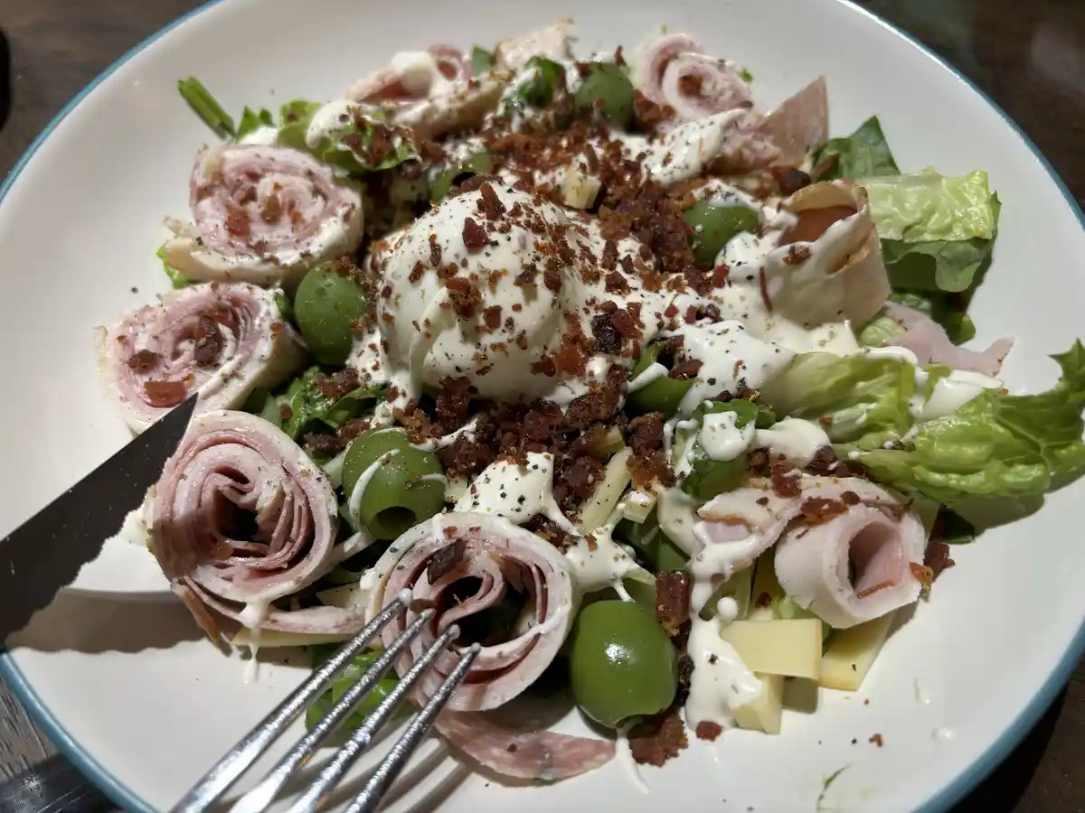 A close-up photo of a white plate with a salad featuring lettuce, rolled ham, green olives, crumbled bacon, cheese, and a…