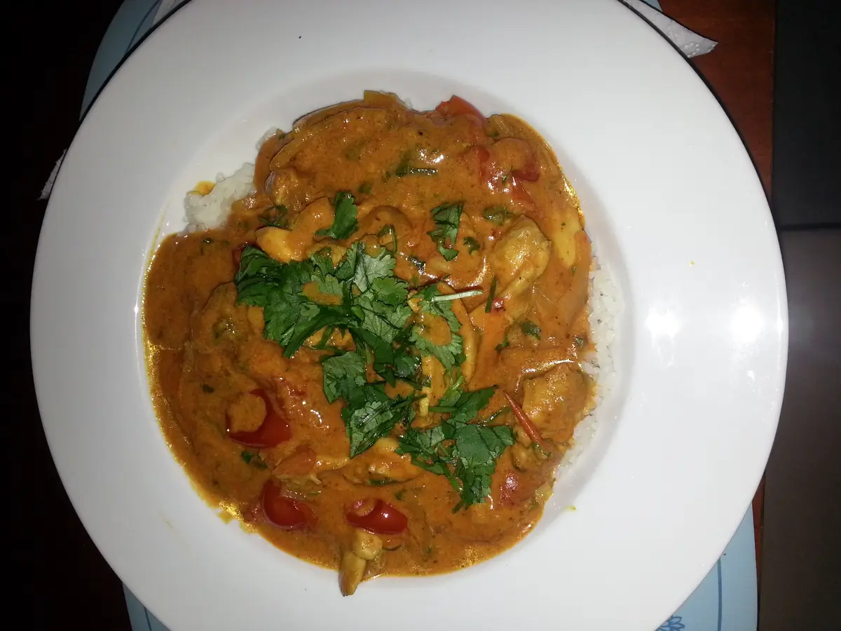 A top-down photo of a white plate holding a dish of orange-colored curry with chunks of meat, red peppers, and fresh…