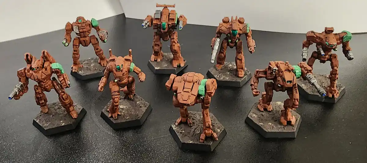 Seven painted miniature mechs, mostly orange with green accents and glowing teal eyes, stand on hexagonal bases on a dark…