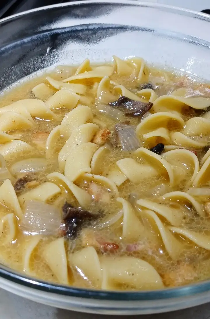 a close-up photo of a glass bowl filled with boiling, yellowish broth containing wide, flat pasta noodles, chunks of meat…