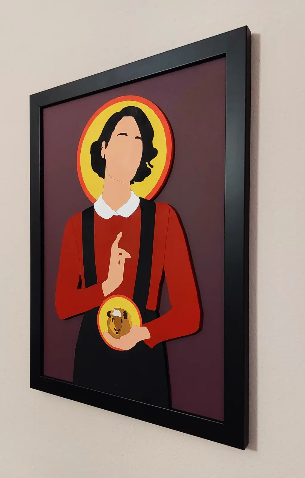 A framed artwork depicts a stylized saint with a halo, wearing a red shirt and black overalls, holding a guinea pig in a…