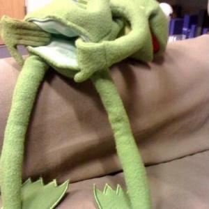 A green plush Kermit the Frog toy is sitting on a brown couch with his legs spread wide apart, appearing to be in a relaxed…