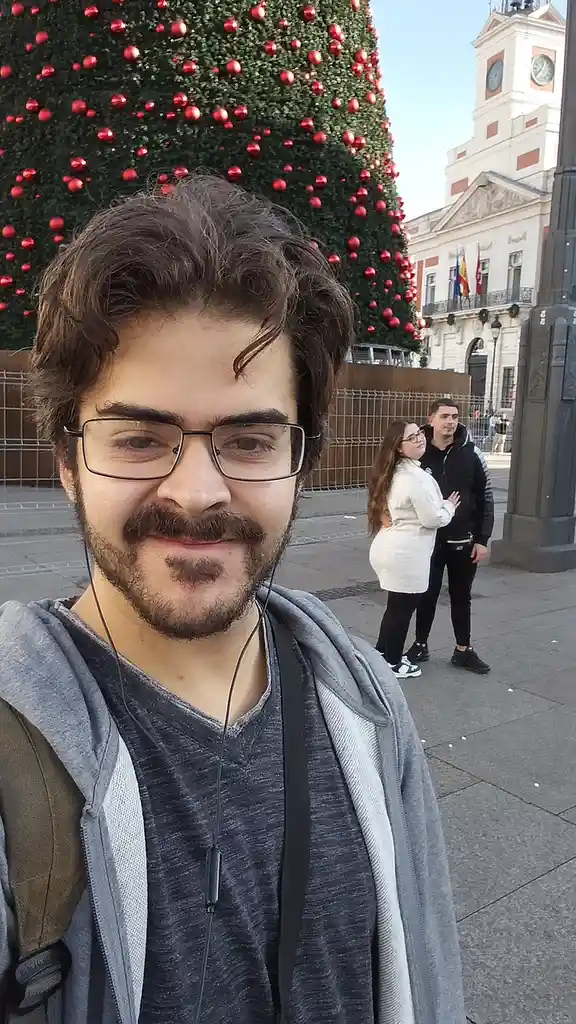 A man with glasses and a beard takes a selfie in front of a large, decorated Christmas tree, smiling at the camera while a…