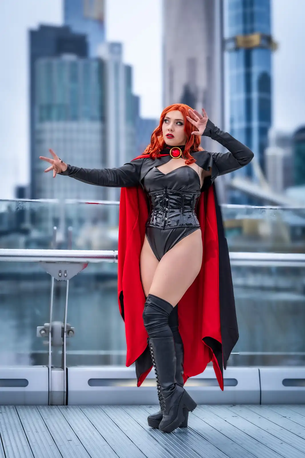 A person in a black and red superhero costume with a cape, corset, and thigh-high boots poses dramatically on a modern city…