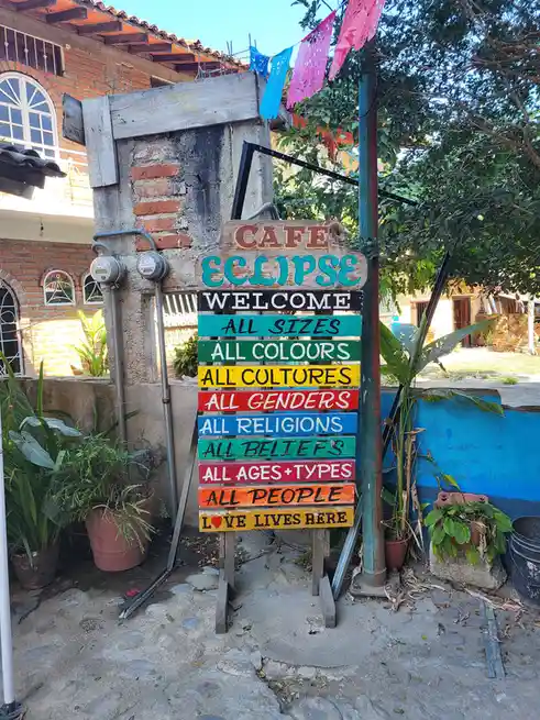 A colorful wooden sign for 'Café Eclipse' stands outdoors, listing welcoming messages like 'All Sizes', 'All Cultures', 'All…