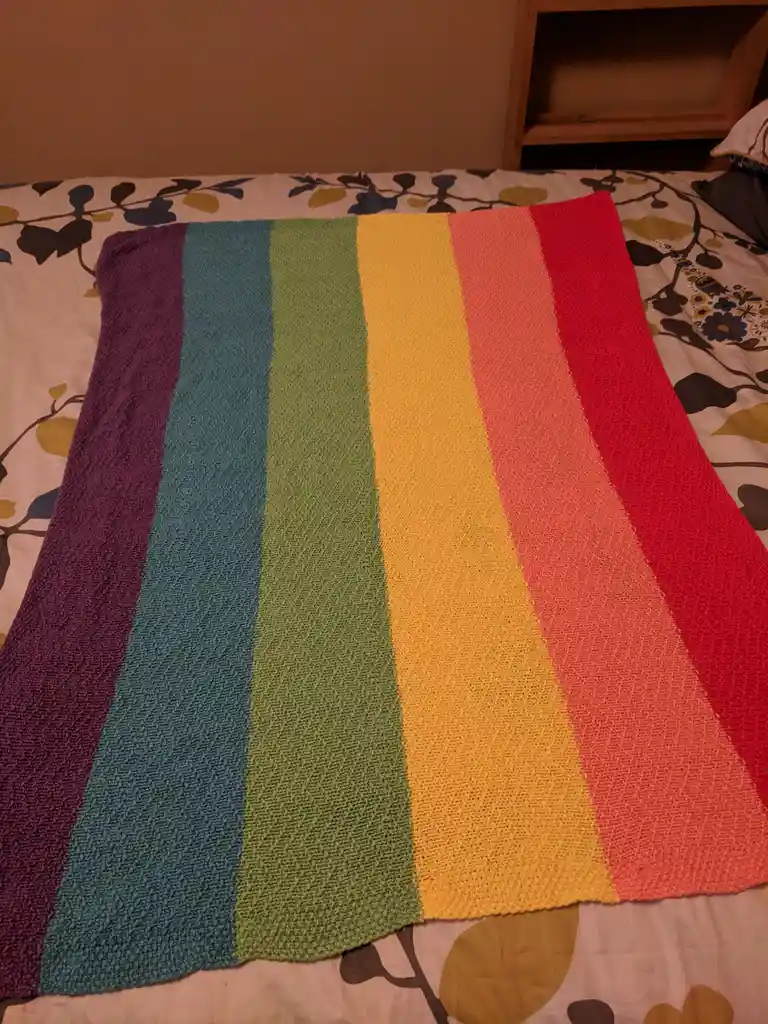 A hand-knitted blanket with vertical rainbow stripes (purple, blue, green, yellow, orange, red) lies on a bed with a…