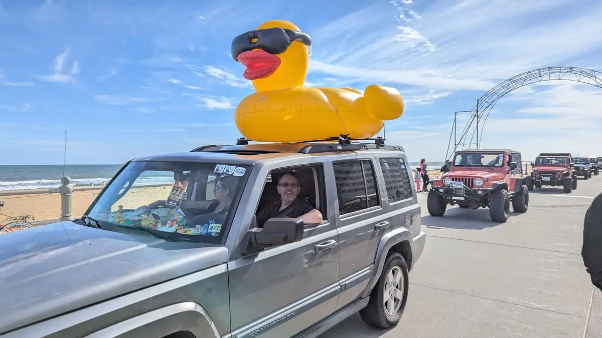 A silver Jeep Commander drives along a beachside road with a giant inflatable yellow rubber duck wearing black sunglasses…