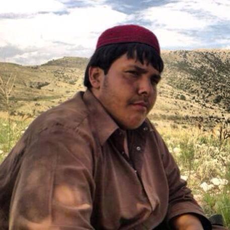 a man wearing a red hat and brown shirt sits in a grassy field with hills behind him, looking directly at the camera with a…