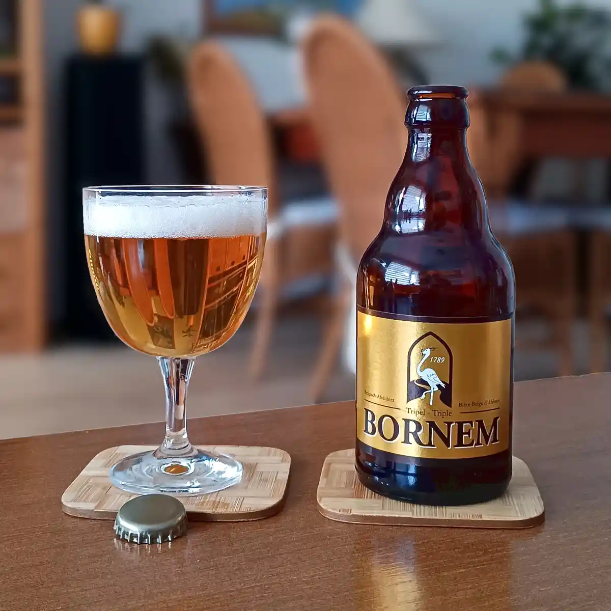 A bottle of Bornem Triple beer and a glass filled with amber beer and white foam sit on wooden coasters on a table, with a…