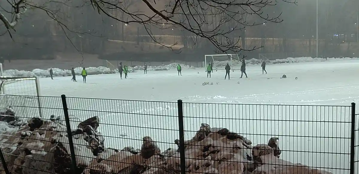 A wide shot of a soccer field completely covered in snow, with several players in winter clothing and bright green vests…