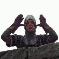 A man in medieval-style armor and a helmet stands on a stone wall, holding his gloved hands up to his ears in a gesture of…