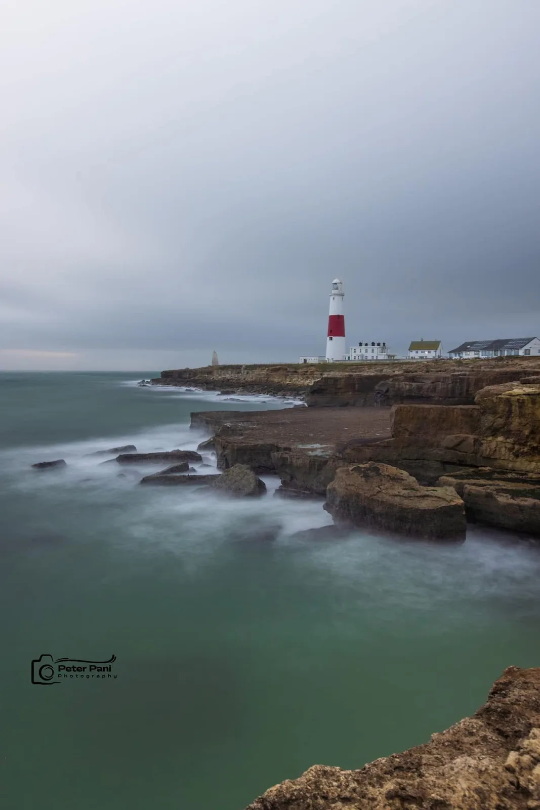 A long-exposure photograph shows a white lighthouse with a red band standing on a rocky cliff overlooking the sea under an…