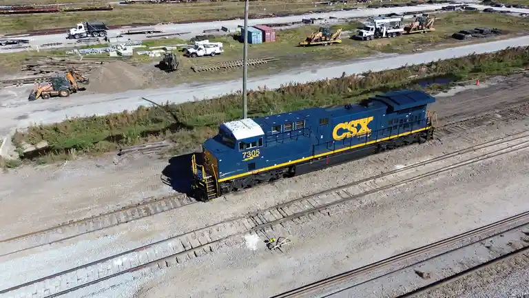 An aerial photo shows a blue and yellow CSX locomotive, number 7305, sitting on railroad tracks in an industrial yard. The…