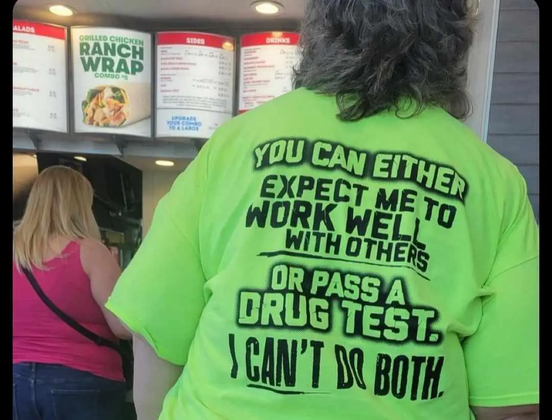 A person with long dark hair wears a bright neon green t-shirt with bold black text on the back, standing at a fast-food…