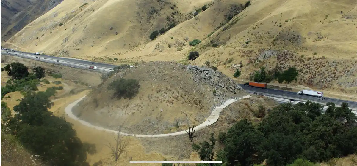 An aerial view of a winding dirt path circling a small hill, with a multi-lane highway running parallel above it, carrying…