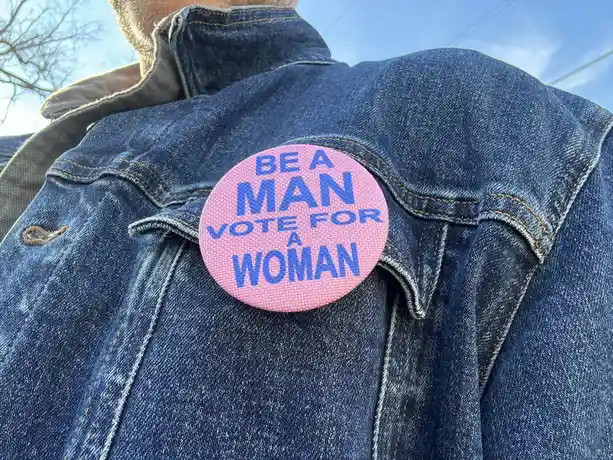 A close-up photo shows a pink circular pin with blue text reading 'BE A MAN VOTE FOR A WOMAN' pinned to the shoulder of a…