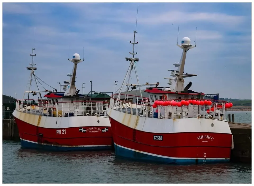 Two red and white fishing boats, 'Amber Mabel' and 'Millie C', are docked side-by-side in a harbor. The boat on the right…