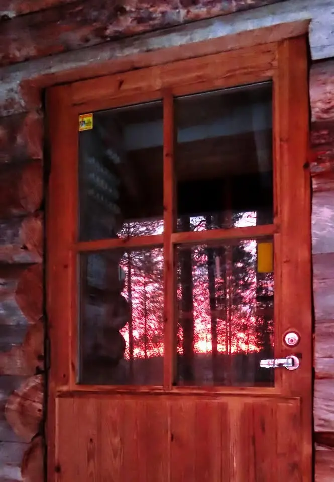 A wooden door with glass panes set in a log cabin wall, showing a reflection of trees bathed in red light, with a yellow…