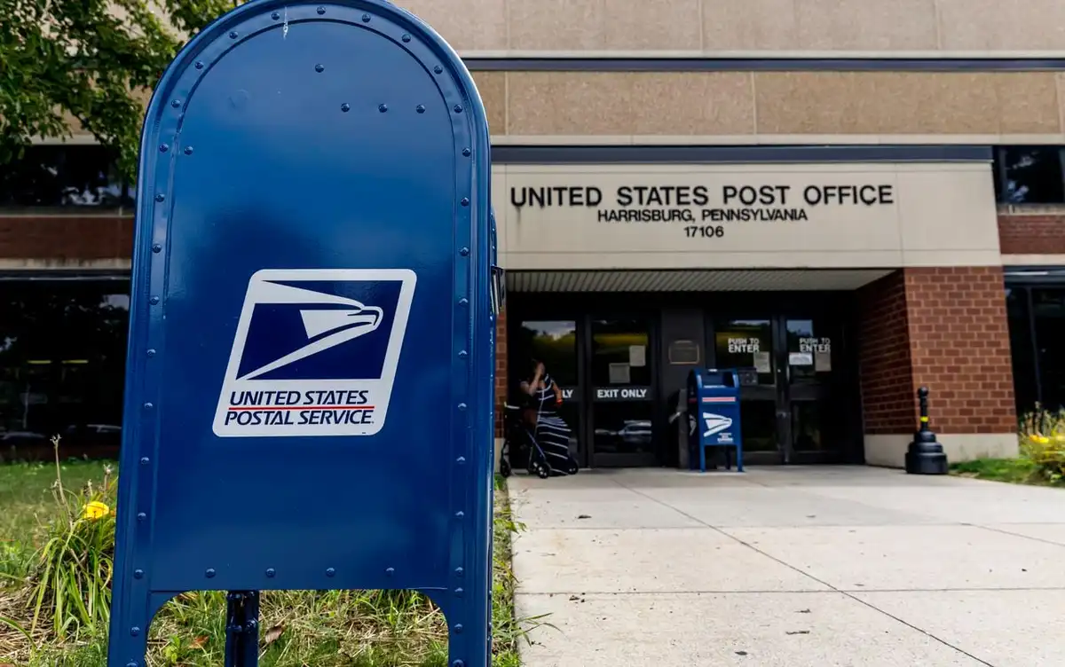 A blue United States Postal Service mailbox stands in the foreground, with a USPS post office building in the background…