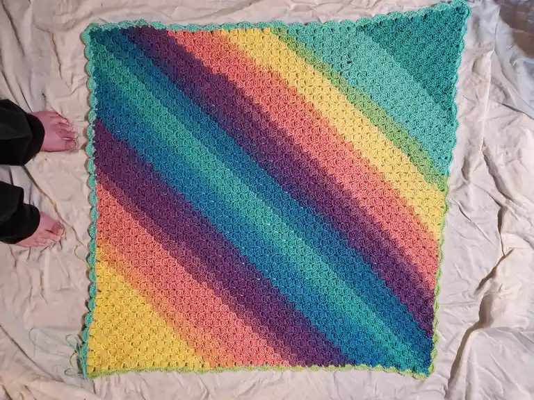 An overhead photo shows a large, colorful crocheted blanket with diagonal rainbow stripes laid out on a wrinkled white…
