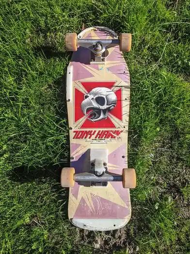 A vintage Tony Hawk skateboard with a skull graphic and red cross design lies on green grass. The board features the text…
