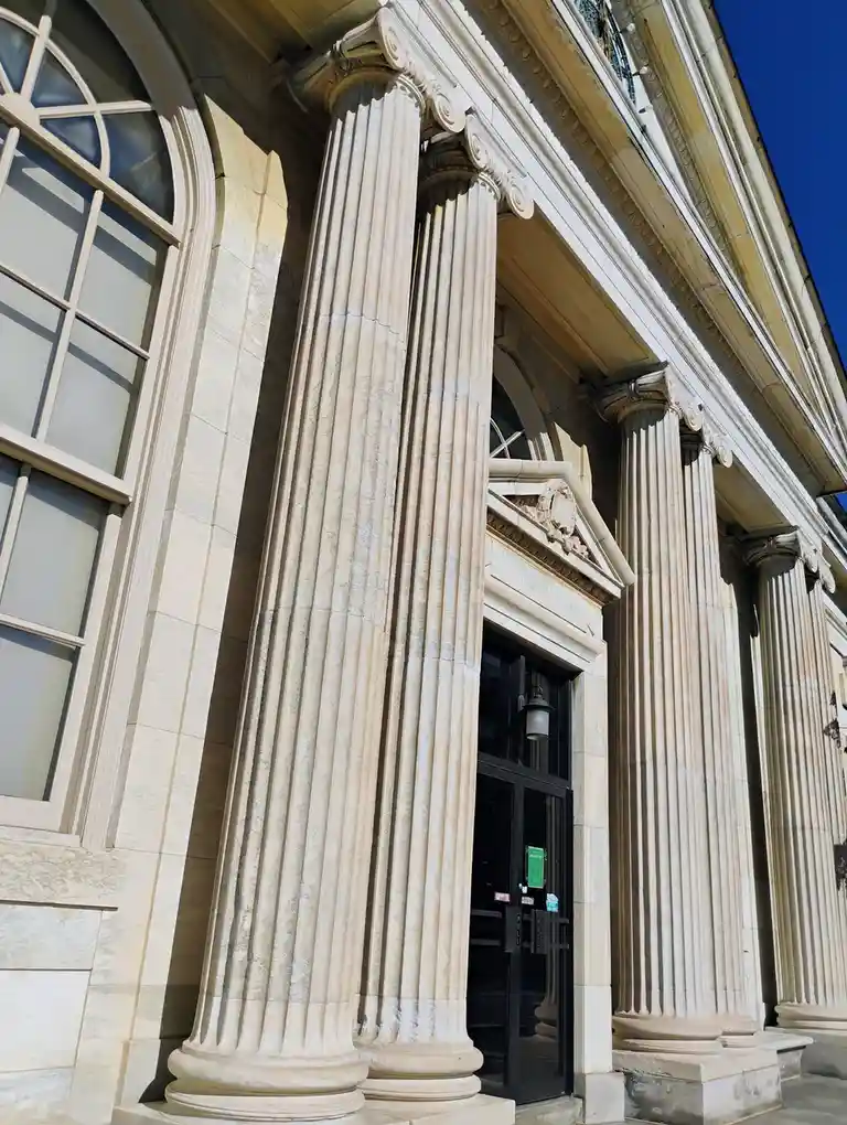 A low-angle photo shows the grand, classical facade of a stone building with large fluted columns and arched windows under a…