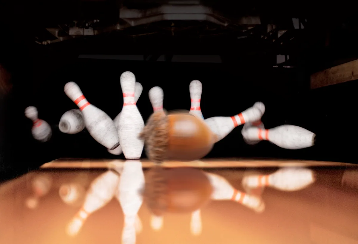 A blurry, low-angle photo captures a bowling ball in motion, striking white bowling pins with red stripes on a lane. The…