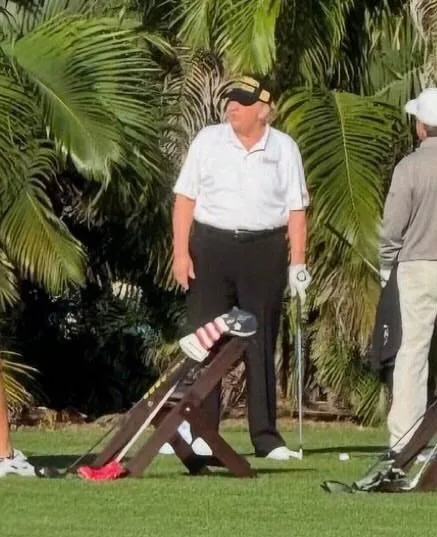 A man in a white polo shirt and black pants stands on a golf course with palm trees behind him, his left shoe is missing and…