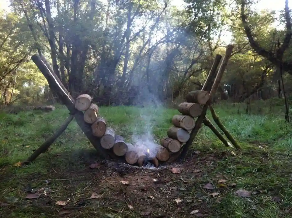 An outdoor scene in a grassy forest with a makeshift hammock constructed from logs arranged in a U-shape, supported by two…