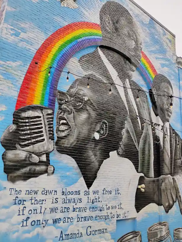 A mural on a blue brick wall depicts three Black historical figures in black and white, with a vibrant rainbow arching over…