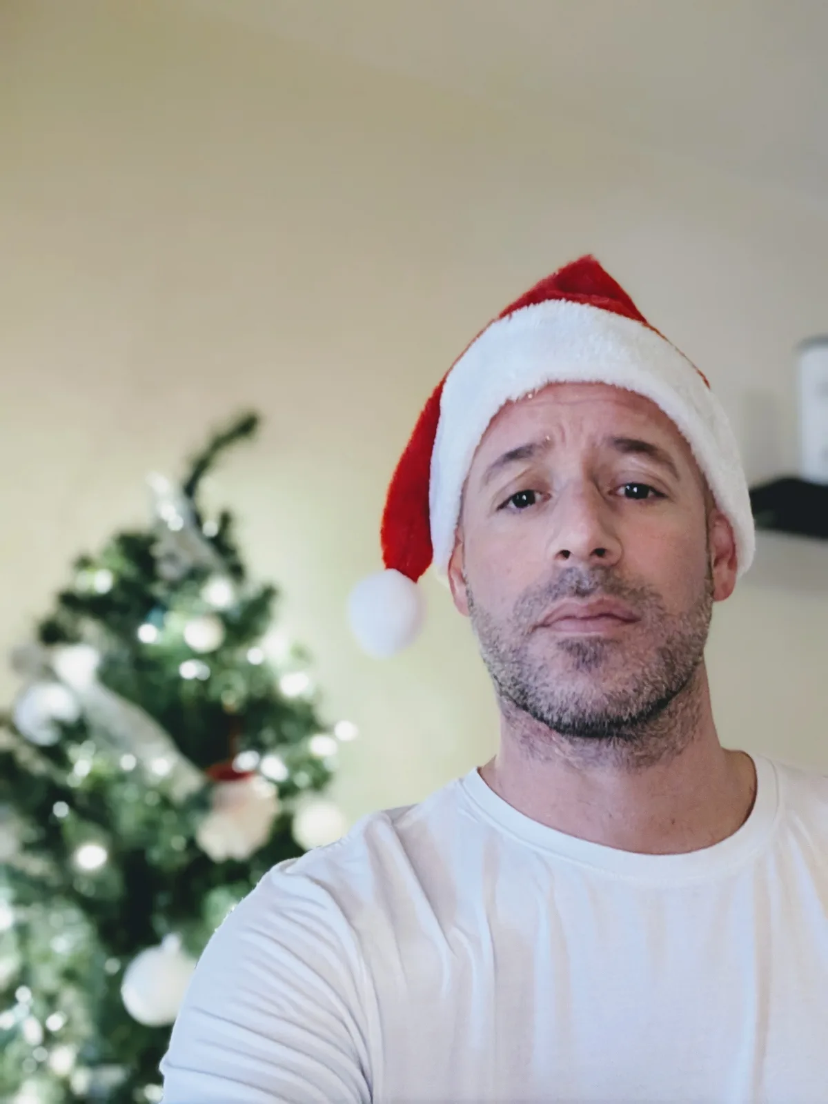 A man with a weary, unenthusiastic expression wears a red and white Santa hat while standing in front of a blurred…